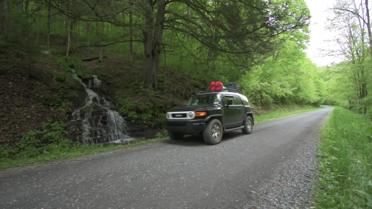 My black Toyota FJ Cruiser with camping gear on the roof driving along a quiet gravel forest road past a small roadside waterfall amid bright spring foliage.