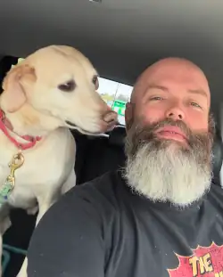 Self portrait of a blue-eyed, bald-headed, bearded man in his car with his yellow lab whippet mix dog, over his shoulder staring in his face.
