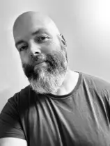 Black and White selfie of a white, cisgender, middle-aged, bald man with a medium-length, salt and pepper beard in a T-Shirt smiling at the camera
