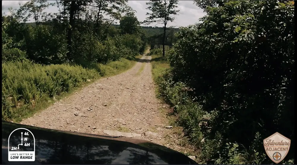 Berry Mountain climb picture 1. View over the hood of the FJ from the hero camera mounted to the windshield. Cruiser hood visible with a dusty-looking dirt and gravel path stretching out before it and green foliage on either side. Sticker on the bottom left of the image that says "Life is better in Low Range"