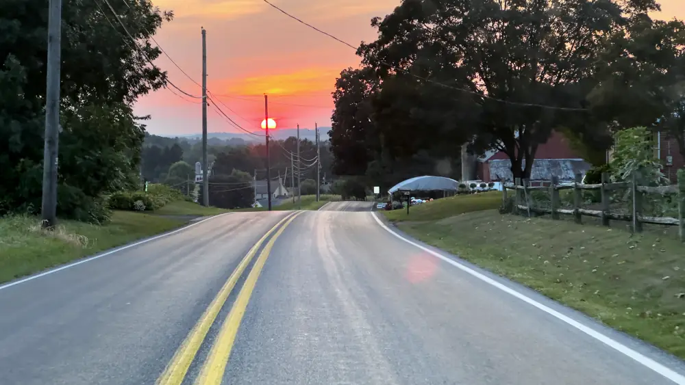 Picture of a road extending off into a sunset with trees, and a barn on the sides of the road.