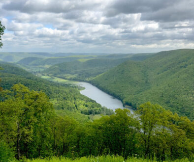 Standing on top of a hill overlooking Kettle Creek State Park. A river is framed by two green-tree covered hills on either side and a partly cloudy sky darkens the image. Just one of many views as a trail guide.