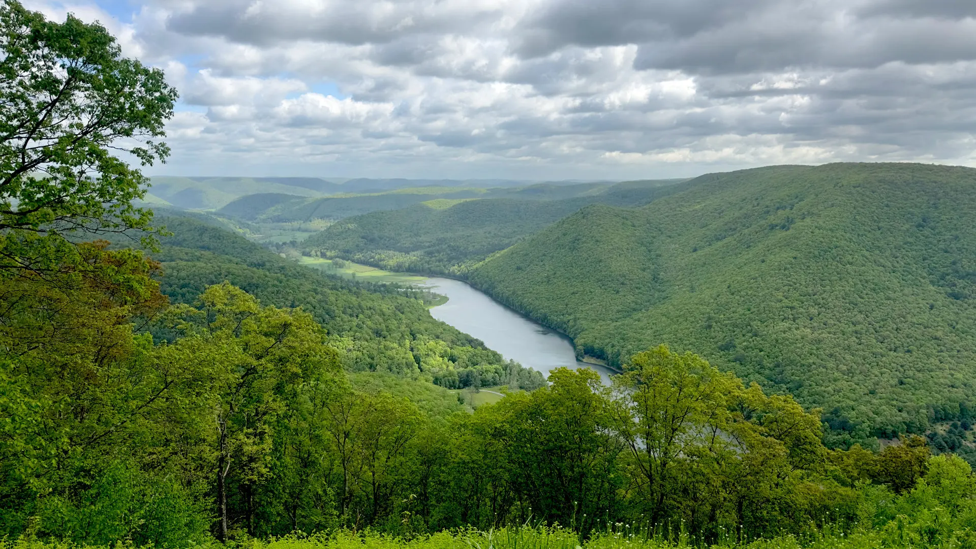 Standing on top of a hill overlooking Kettle Creek State Park. A river is framed by two green-tree covered hills on either side and a partly cloudy sky darkens the image. Just one of many views as a trail guide.
