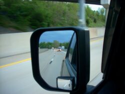 An in-motion shot in the rearview. Three cars approach from behind, and three tractor trailers are parked behind a barrier. Lush spring greens dominate the motion-blurred surroundings of the mirror.