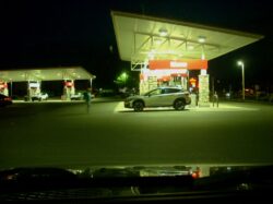 A shot out the windshield of a parked car at a Wawa gas station just after dusk. A Subaru waits patiently at the nearest gas pump as its motion blurred owner walks back towards it. More cars can be seen parked at pumps, moving near pump, and moving down the street in the background.