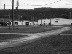 Black and white photo of two buildings at the Bloomsburg Fair Grounds from across a rain-soaked parking area. One building says "Cattle" the other says "Arena"