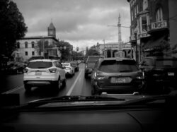 A black and white image through the windshield of a car in light Saturday traffic on Main street at Market street in Bloomsburg, PA. The cloud cover is thick but fluffy and well defined. A clock tower rises from a building in the distance on the left and a Column with statues rises from the middle of Market street on the right. Art Deco-style rows of buildings line the right side of the Main before market. More modern buildings are on the other side of Market.