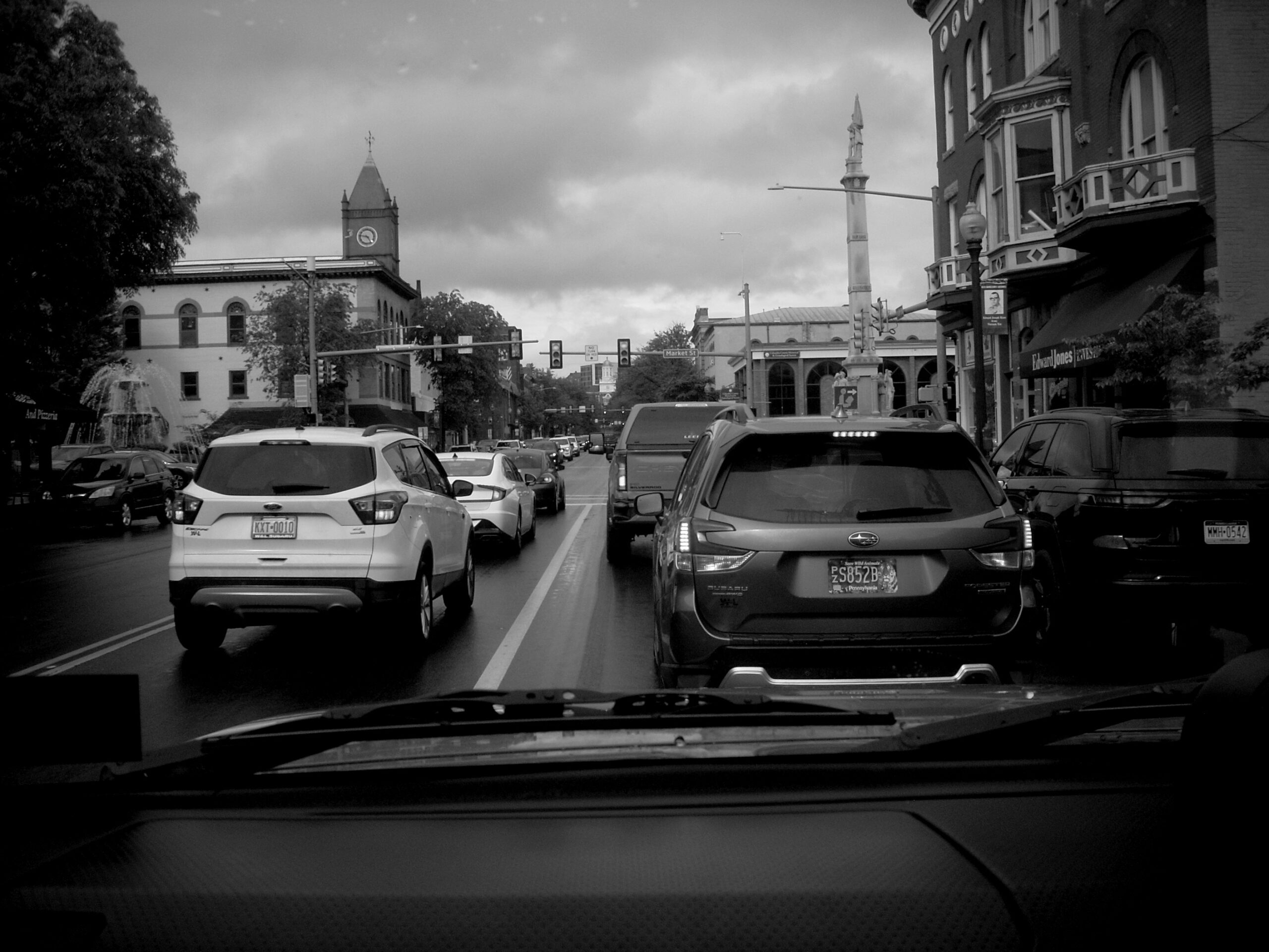 DSCN0417 A black and white image through the windshield of a car in light Saturday traffic on Main street at Market street in Bloomsburg, PA. The cloud cover is thick but fluffy and well defined. A clock tower rises from a building in the distance on the left and a Column with statues rises from the middle of Market street on the right. Art Deco-style rows of buildings line the right side of the Main before market. More modern buildings are on the other side of Market.