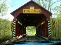 Photo through the windshield as I approach a covered bridge framed by trees near Elysburg, PA, home of Knoebel's amusement park. A tree-covered hill can be seen in the background and through the bridge a gravel road can be seen. Signs on the bridge read 
